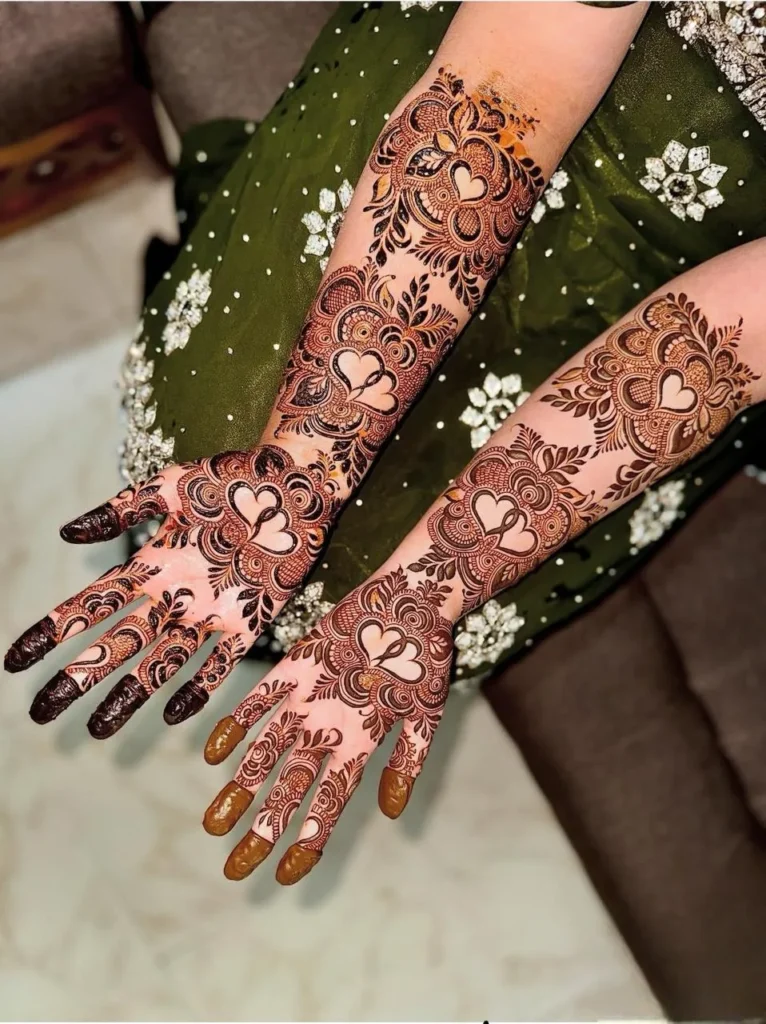 Full-hand bridal mehndi with intricate heart-themed motifs and traditional henna detailing, beautifully showcased against a green embroidered dress